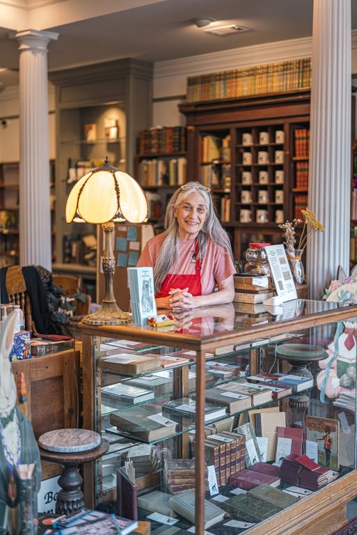 Smiling woman with long gray hair standing behind a glass case of vintage children's books