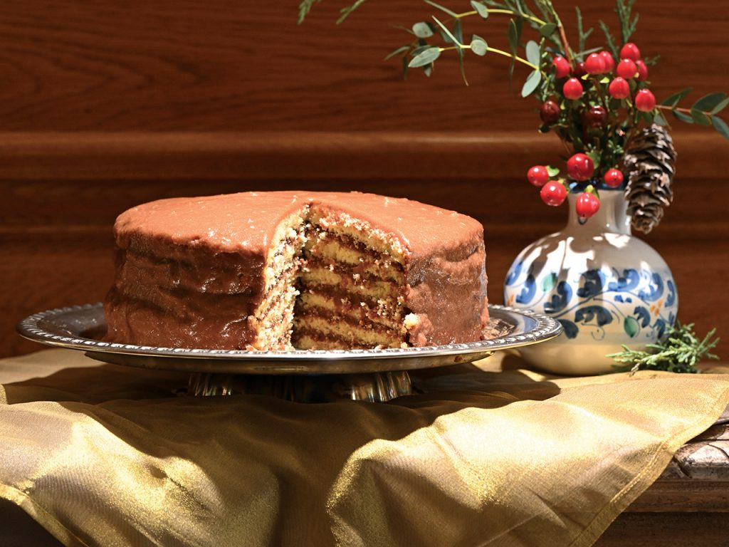 Chocolate layer cake with a piece removed, sitting on a silver platter on a wooden table covered with a gold tablecloth