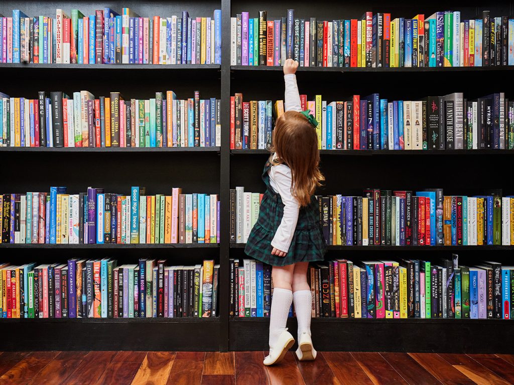 Little girl reaching for a book high on a book shelf