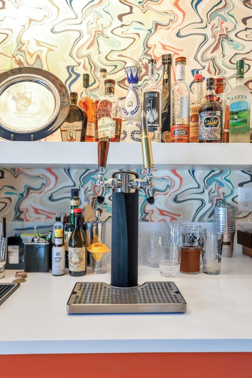 A beer tap in a wet bar with bottles on shelves and funky wallpaper