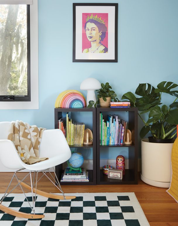 Child's bedroom with pale blue painted walls, shelving full of books and toys, a minimalist white rocking chair and a black and white checkered rug covering the light natural wood floor