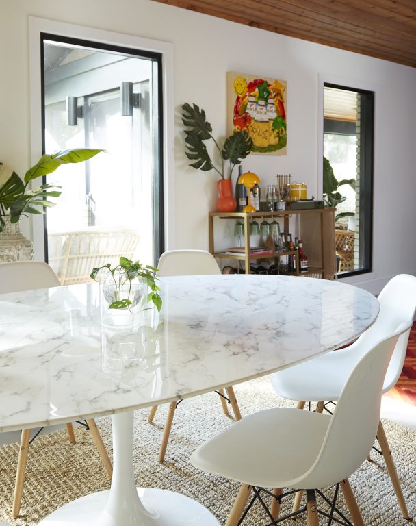 Marble finish dining table with minimalist white plastic chairs in front of a bar cart and a bright colorful painting