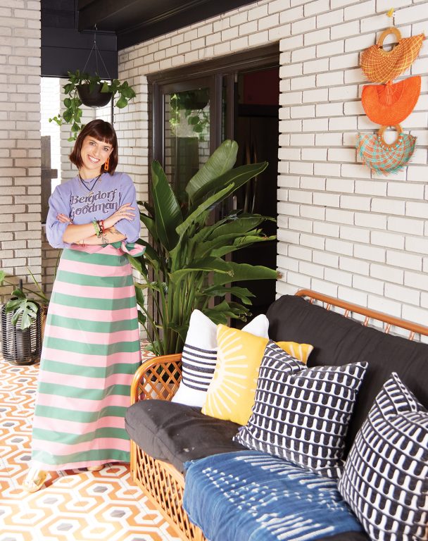 Woman in a green and pink striped long skirt and purple shirt standing on a screened in patio next to a rattan couch