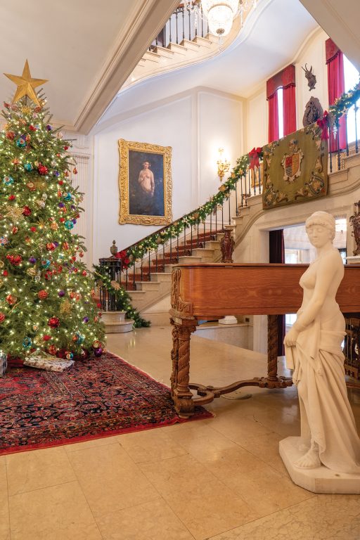 A marble statue of a woman and a grand piano share space with an enormous, brightly lit Christmas tree.