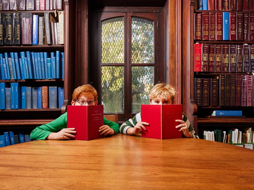 Two boys sitting at a big round table in front of bookshelves and peeking out from behind books