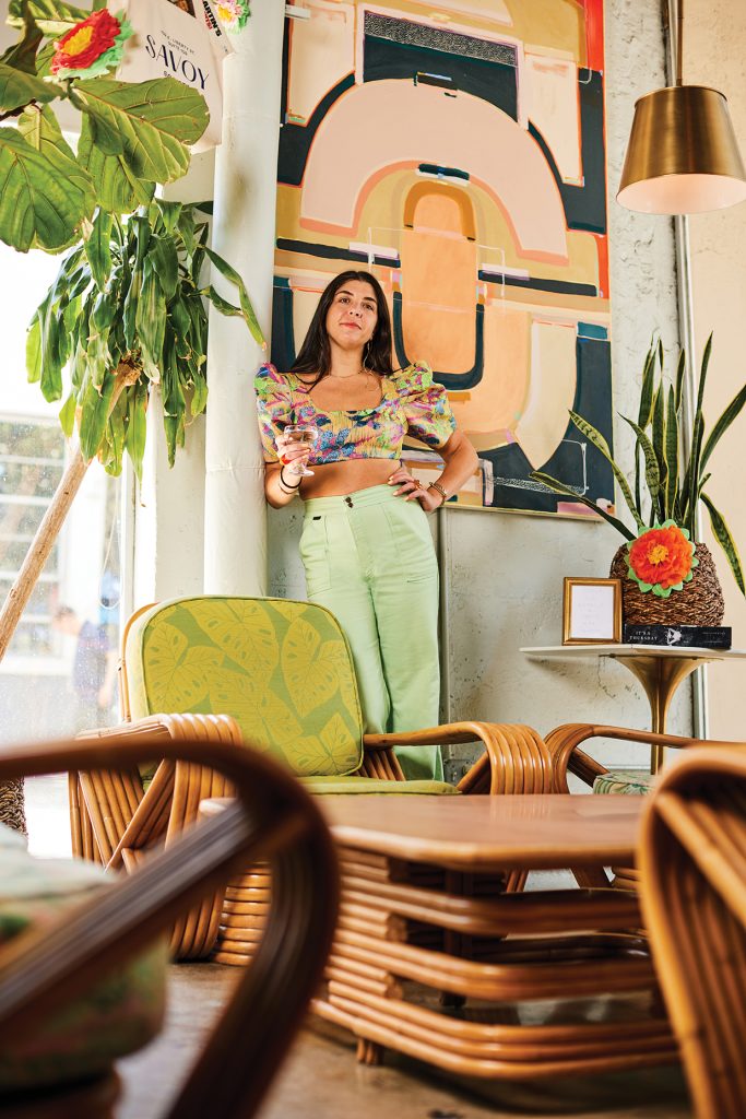 Woman in colorful crop top and mint green pants holding a drink and smiling